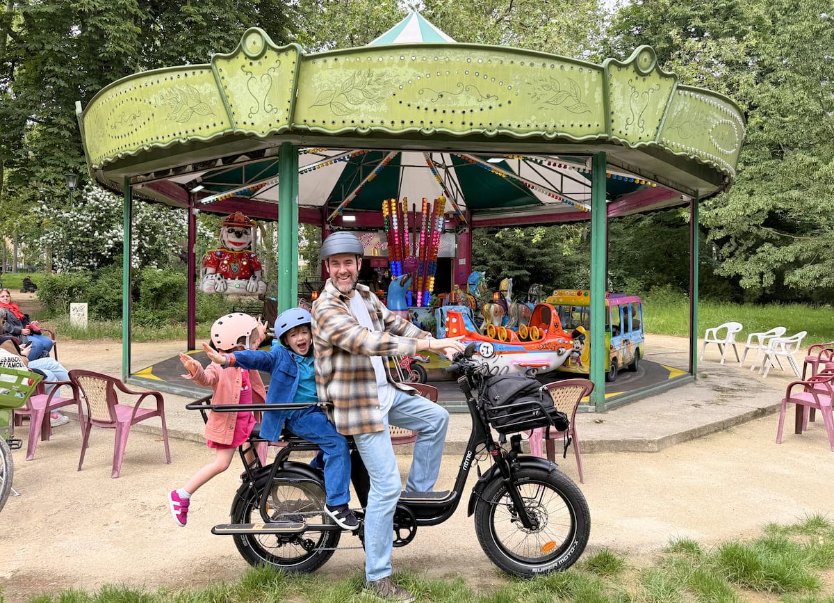 Un adulte et deux enfants sur un vélo cargo Ritmic Jumbo devant un manège, lors d’une sortie en famille à Paris.